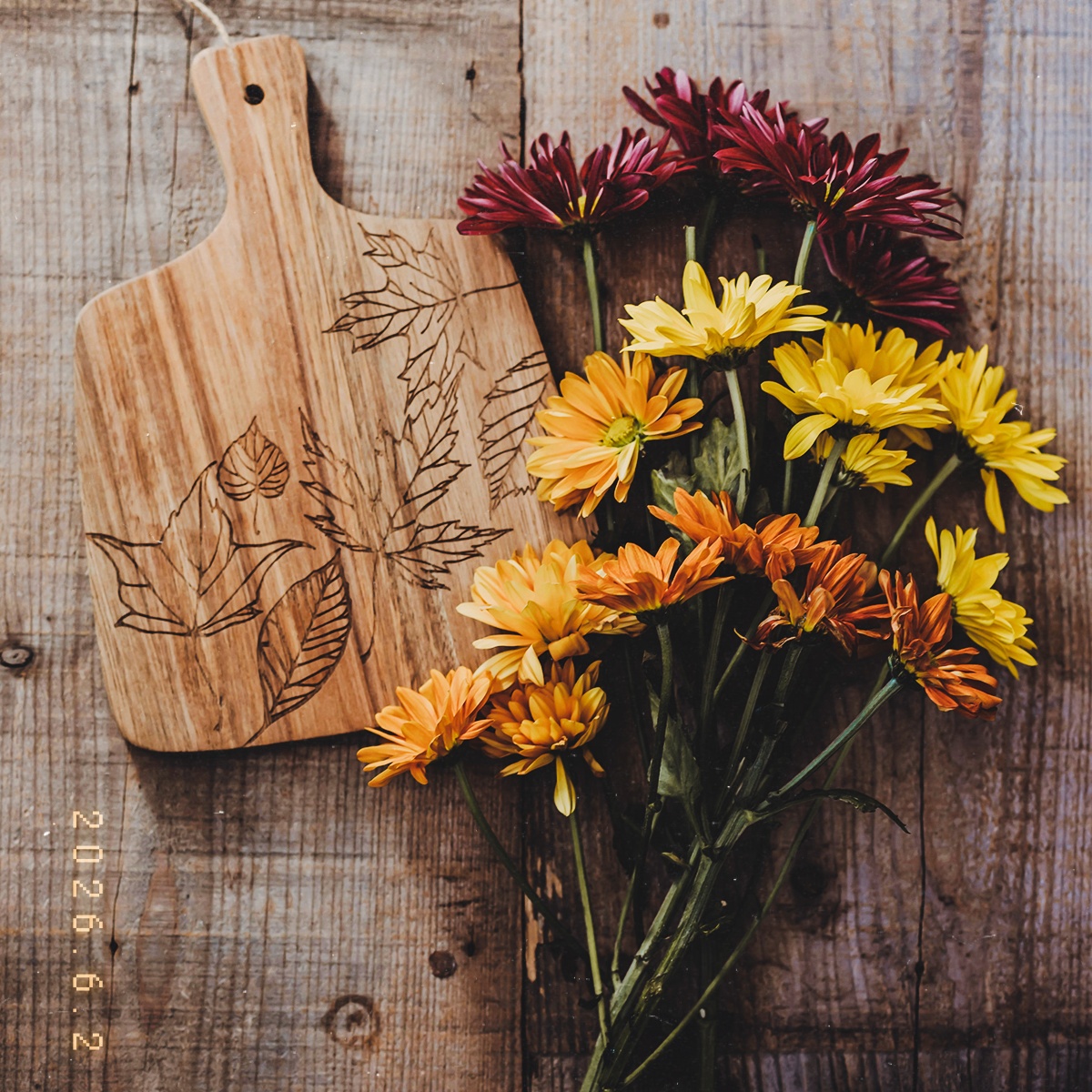 Orange and yellow flowers beside a floral wood-burned cutting board on a rustic table.