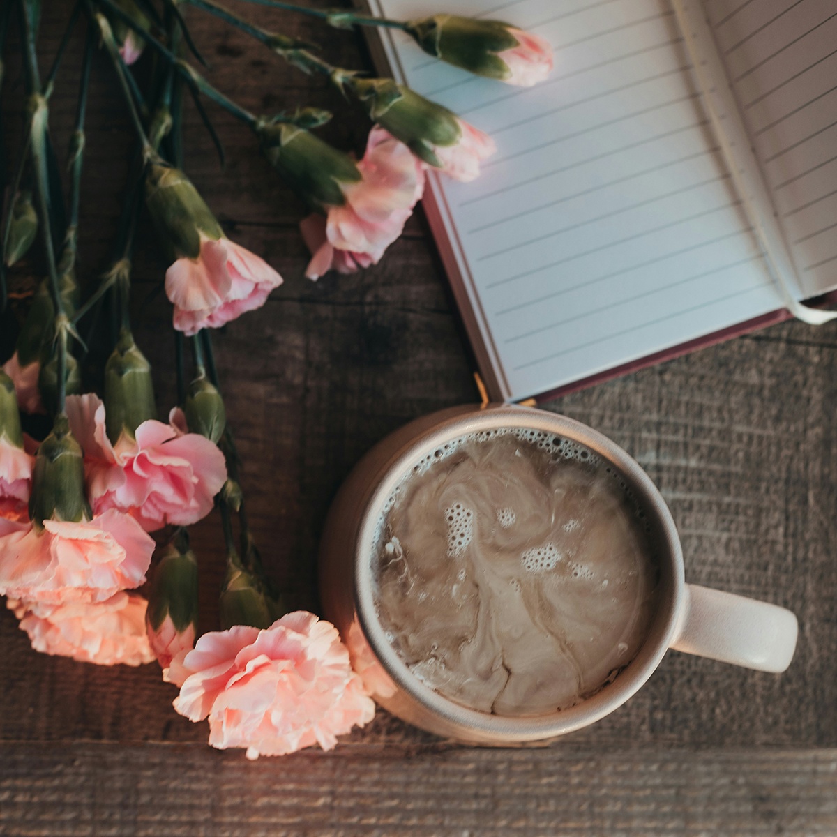 Coffee, pink flowers, and an open notebook arranged on a rustic wooden table.
